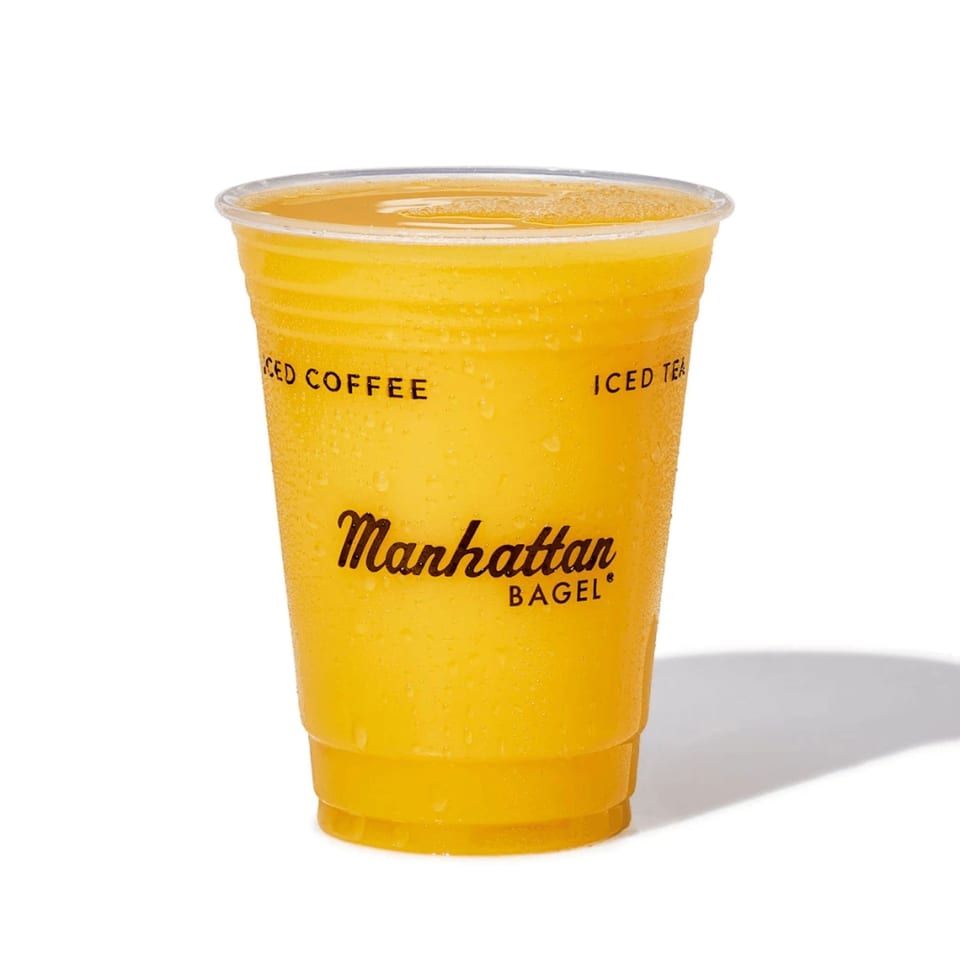 A Manhattan Bagel plastic cup filled with a cold, yellow beverage, likely orange juice.