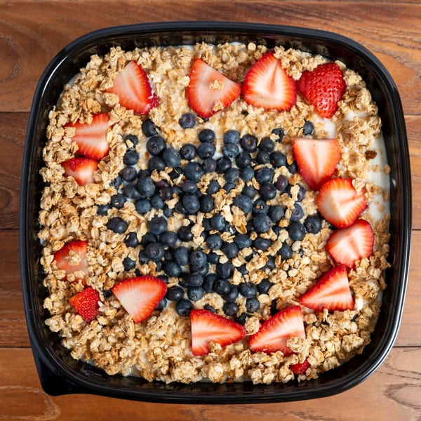 Overhead view of a yogurt parfait with granola, blueberries, and sliced strawberries in a black container.