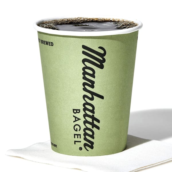 A green Manhattan Bagel coffee cup filled with dark coffee and foam, resting on a white napkin.