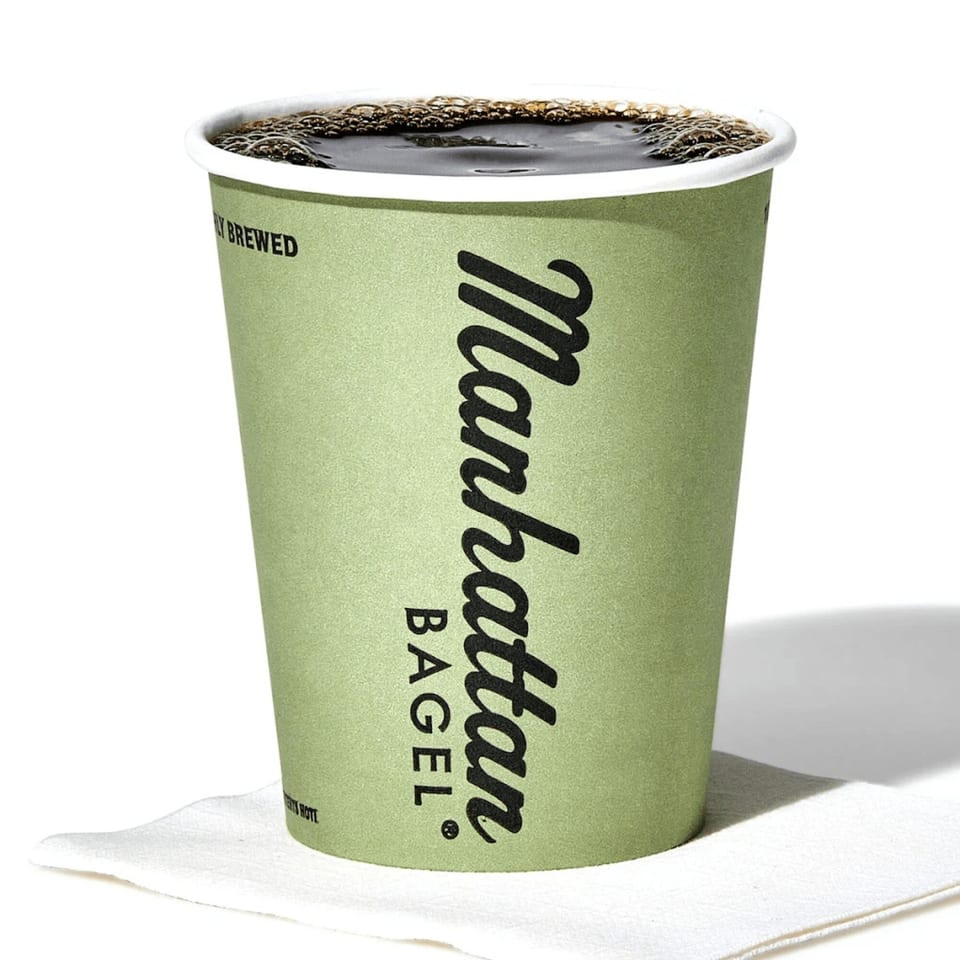 A green Manhattan Bagel coffee cup filled with dark coffee and foam, resting on a white napkin.