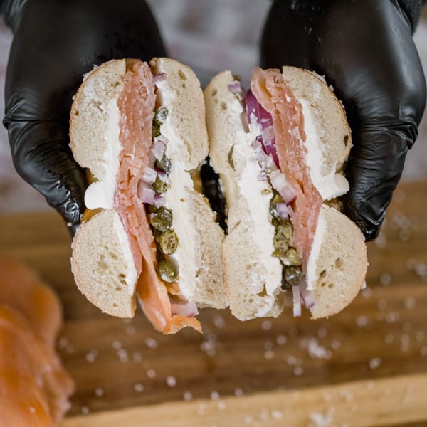 Hands in black gloves hold a sliced bagel filled with lox, cream cheese, red onion, and capers.