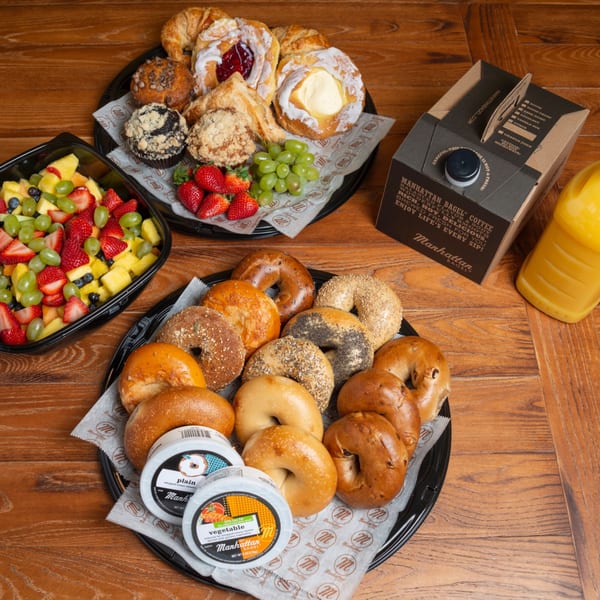 A Manhattan Bagel catering spread with bagels, pastries, fruit salad, coffee box, and orange juice.