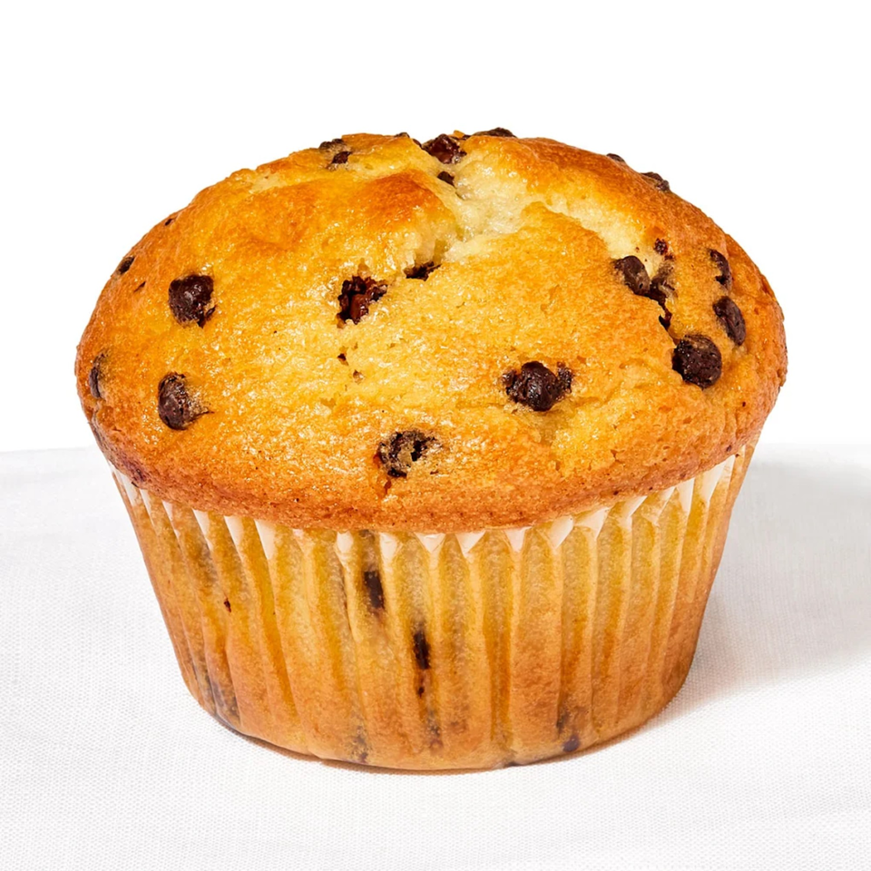 A golden-brown chocolate chip muffin in a paper liner sits on a white surface.